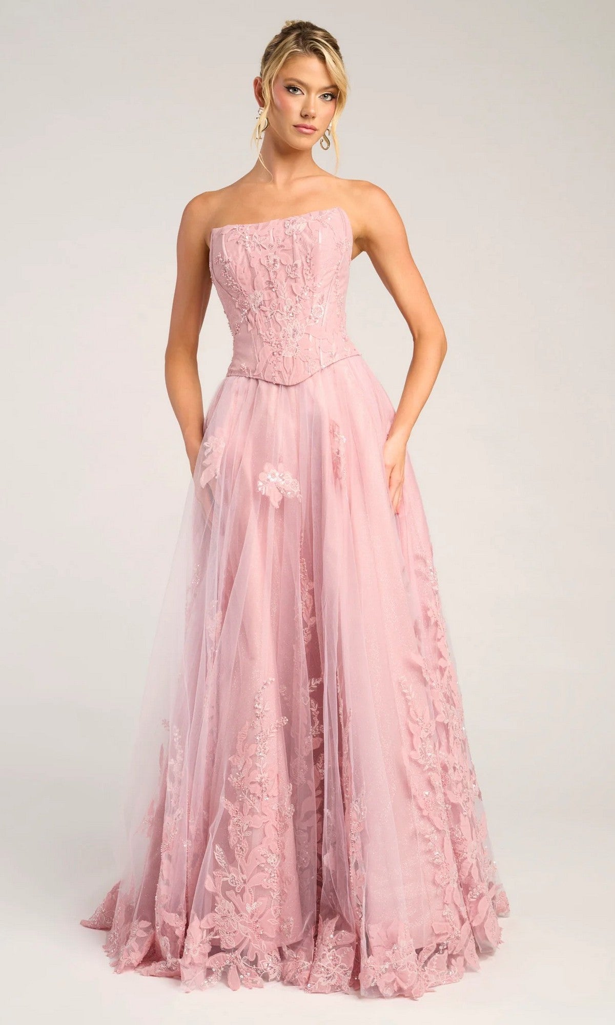 A woman in the Portia and Scarlett PS26061 long formal pink dress, featuring a sleeveless design, floor-length skirt, corset bodice, and floral lace details, stands against a plain light background.